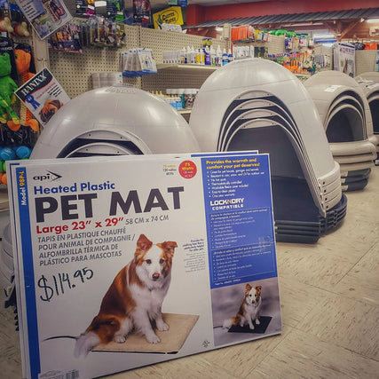 PetStore aisle filled with stacked pet igloos. Foreground shows a large sign for a heated plastic pet mat with an image of a dog, priced at $114.95.
